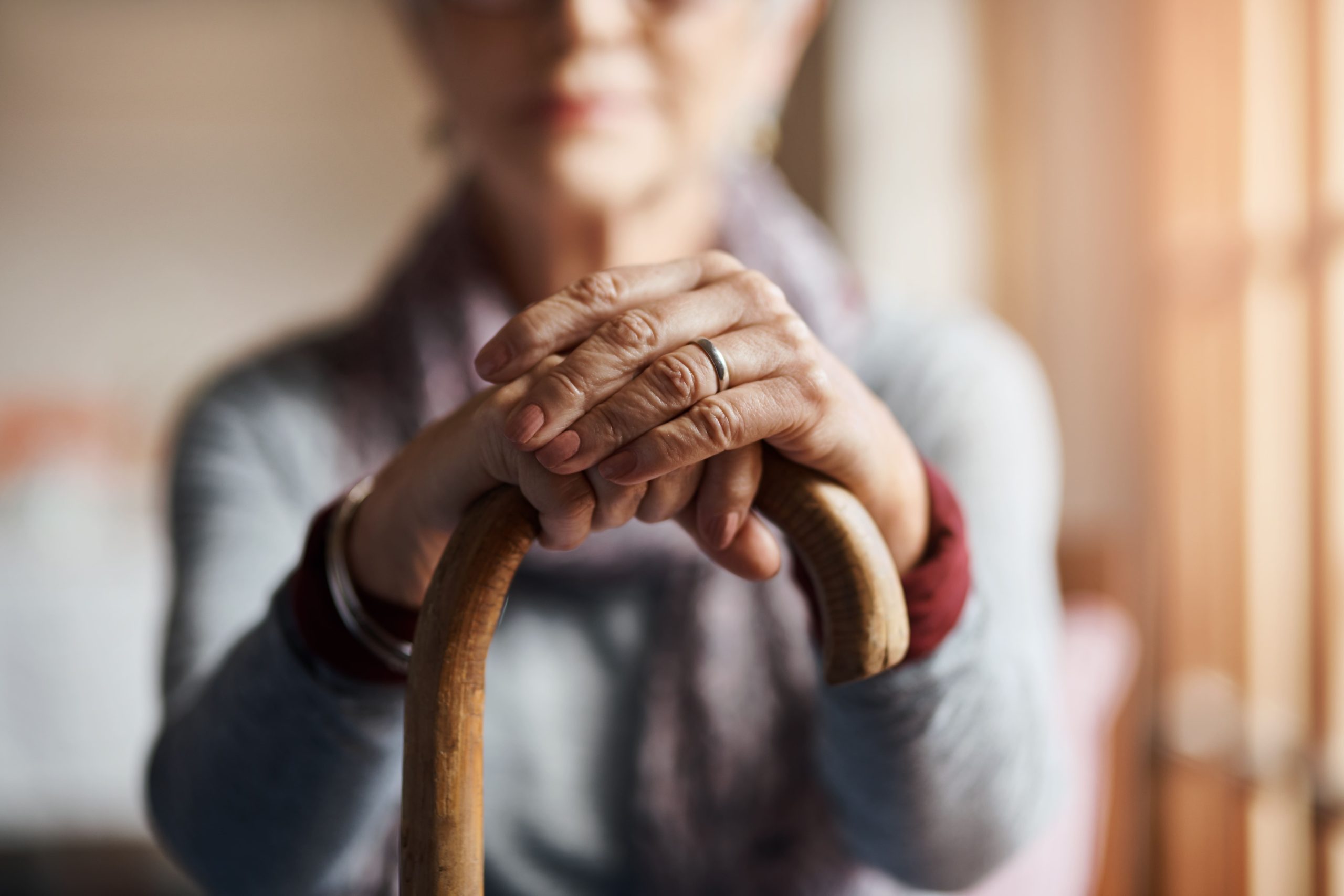 An elderly woman sitting down while holding a cane out in front of herself, both hands are resting on top of the cane.