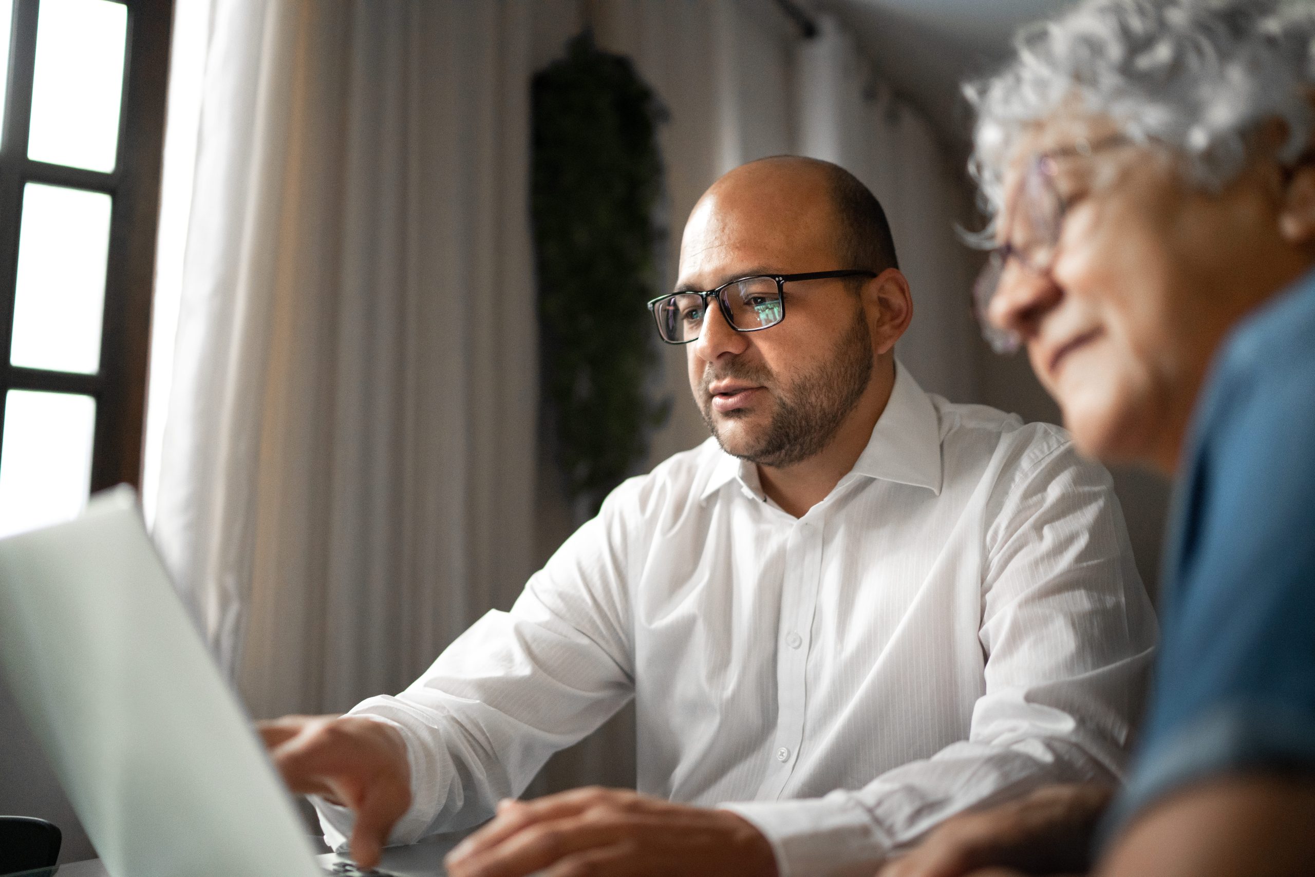 A middle-aged man shows an elderly woman something on a laptop while they sit at a table together.