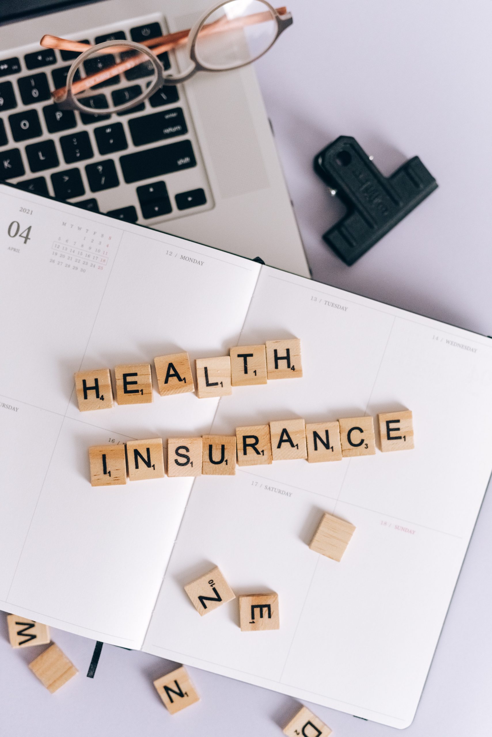 A daily planner is open on a laptop on a table with scattered Scrabble tiles. Some of the Scrabble tiles spell out "Health Insurance".