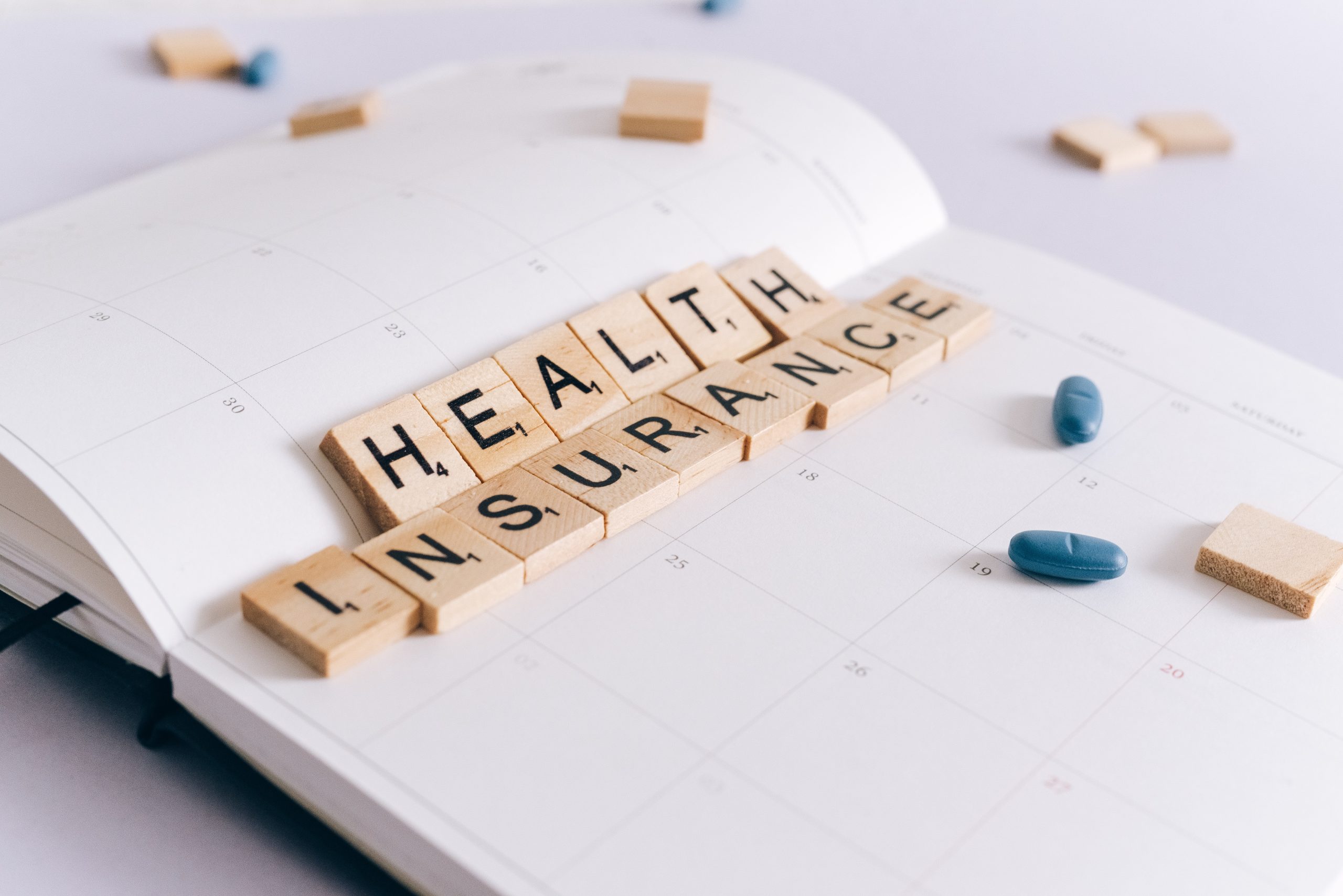 A daily planner is open on a table with scattered Scrabble tiles and prescription medication. Some of the Scrabble tiles spell out "Health Insurance".