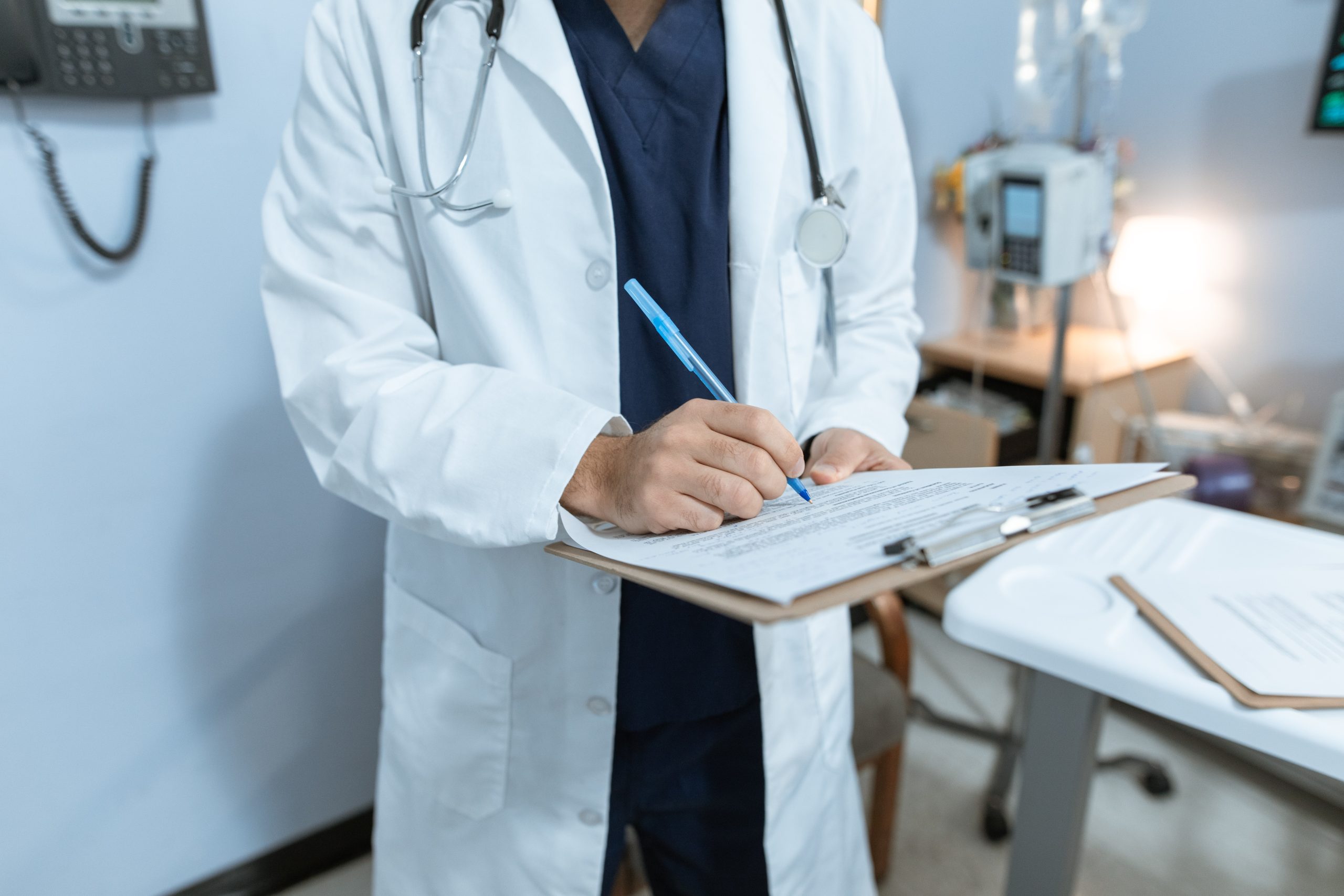 A person in a white doctor coat with a stethoscope around their neck fills out paperwork on a clipboard in a clinic setting.